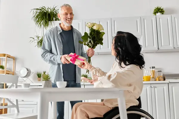A heartwarming scene unfolds as a man affectionately gives a bouquet of flowers to his wife in a wheelchair in their kitchen at home. — Photo de stock