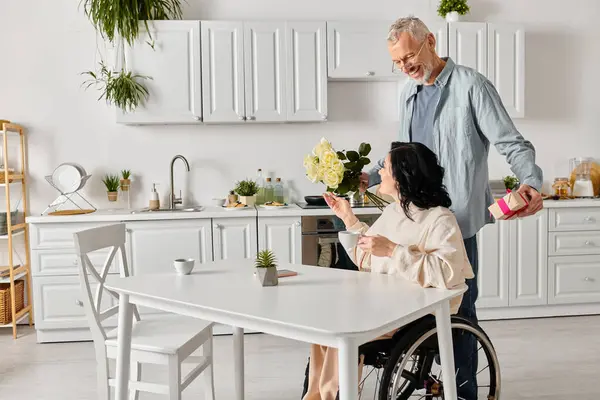 Homme donne affectueusement un bouquet de fleurs à sa femme dans un fauteuil roulant dans leur cuisine à la maison. — Photo de stock