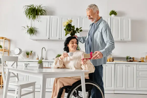 A man standing devotedly next to his disabled wife in a wheelchair in their kitchen at home. - foto de stock