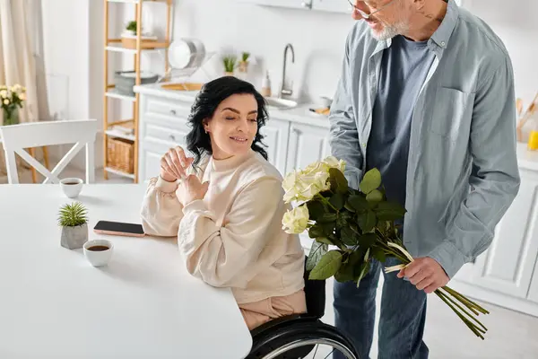 Un homme tenant un bouquet, se tient amoureusement à côté de sa femme alors qu'elle est assise dans un fauteuil roulant dans leur cuisine à la maison. — Photo de stock