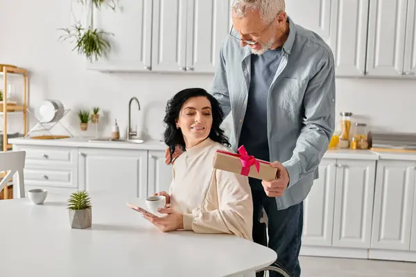 A man lovingly presents a gift to his disabled wife in a wheelchair at a cozy home kitchen table. — Stock Photo