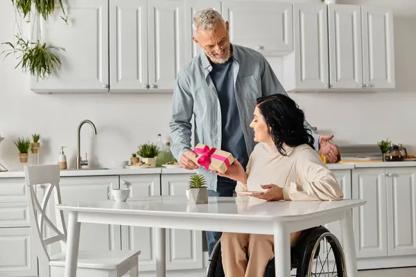 A man tenderly hands a gift to a disabled woman in a wheelchair, in their kitchen at home. — Stock Photo