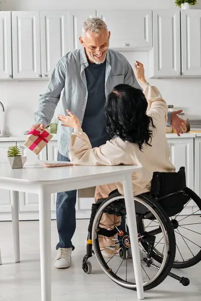 Un uomo fa un regalo a sua moglie su una sedia a rotelle, nella cucina della loro casa. — Foto stock