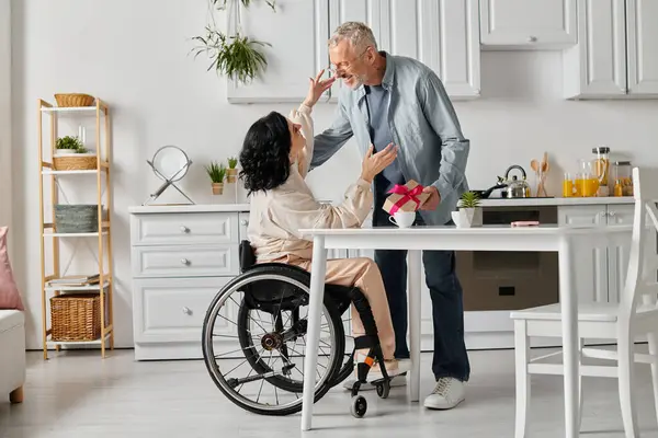 A loving man gives a gift to his happy wife in a wheelchair, in the kitchen of their home. — Stock Photo