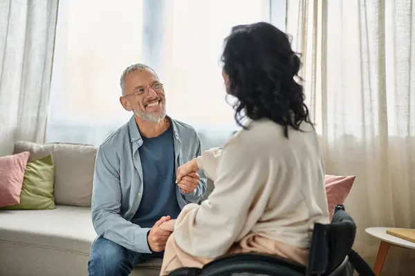 A woman in a wheelchair engaging in conversation with a man, both smiling in a cozy living room setting. — Stock Photo