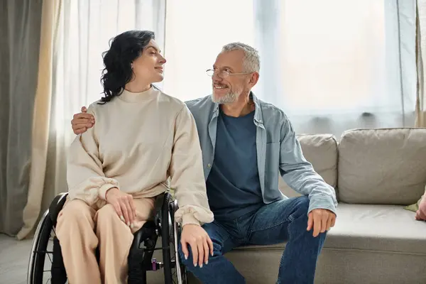 Una mujer discapacitada en silla de ruedas conversa con un hombre en la sala de estar. - foto de stock