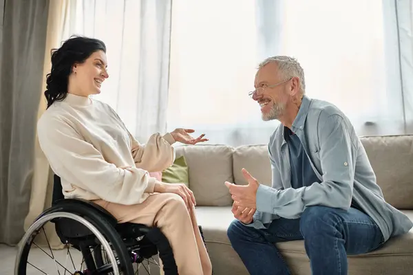 Un hombre conversa con una mujer discapacitada en silla de ruedas en una acogedora sala de estar. - foto de stock