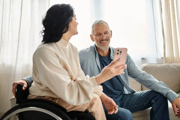 Una donna disabile su una sedia a rotelle tiene un telefono cellulare mentre è seduta nel suo salotto, suo marito al suo fianco. — Foto stock
