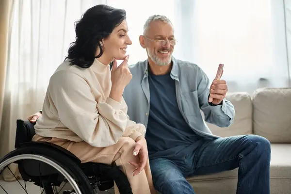 Um homem sentado ao lado de uma esposa com uma deficiência e usando smartphone em um ambiente acolhedor sala de estar. — Fotografia de Stock