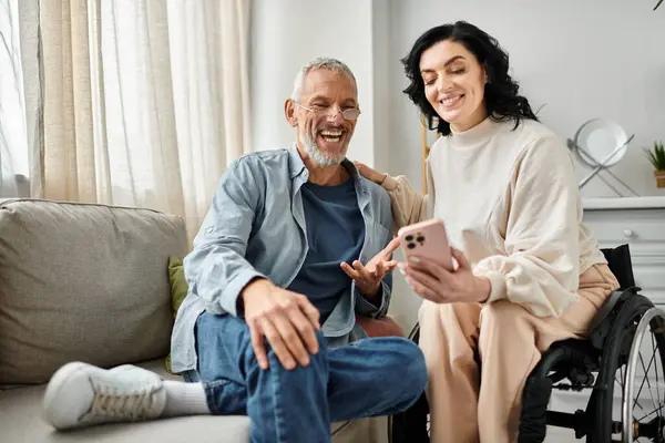 A man sits next to his wife on a wheelchair with smartphone in a cozy living room. — Stock Photo