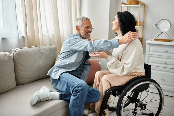 Una donna disabile su una sedia a rotelle sta abbracciando suo marito in modo premuroso e solidale nel loro salotto.. — Foto stock