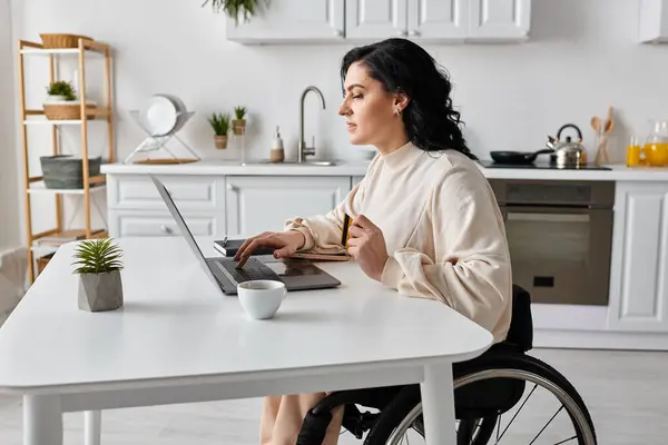 Disabled woman in a wheelchair working on a laptop in her kitchen. — Stock Photo