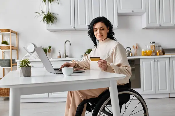 Una mujer discapacitada en silla de ruedas trabaja remotamente en su portátil desde su cocina. - foto de stock
