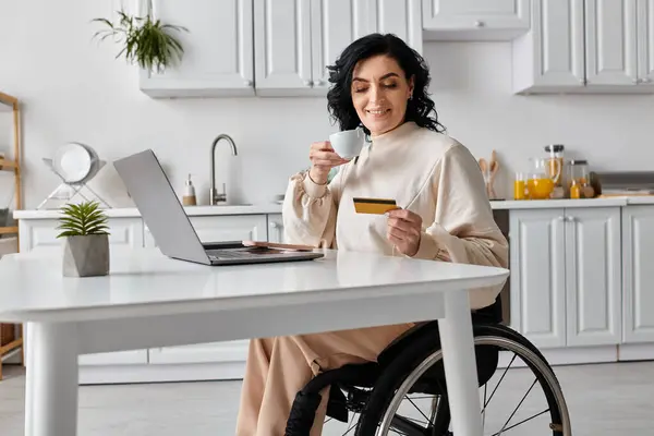 Uma mulher deficiente em uma cadeira de rodas segura uma xícara de café enquanto olha para um cartão de crédito, trabalhando remotamente de sua cozinha. — Fotografia de Stock