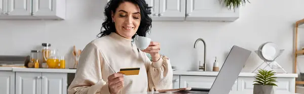 A happy woman holds a credit card while focused on her laptop in the kitchen. — Stock Photo