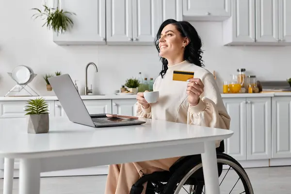 Eine Frau im Rollstuhl kauft selbstbewusst mit Kreditkarte und Laptop in ihrer Küche online ein. — Stockfoto