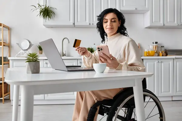 Mujer en silla de ruedas manejando finanzas y trabajando en línea usando un portátil en su cocina. - foto de stock