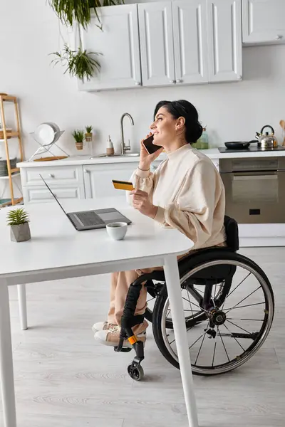 Una mujer discapacitada en silla de ruedas hablando en un teléfono celular mientras trabaja de forma remota desde su cocina. - foto de stock