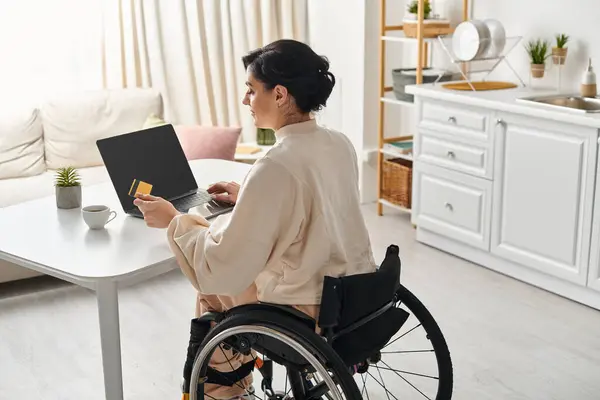 Uma mulher deficiente em uma cadeira de rodas está trabalhando remotamente em seu laptop na cozinha. — Fotografia de Stock