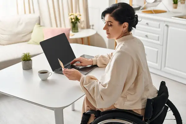 Une femme en fauteuil roulant utilise un ordinateur portable dans sa cuisine, s'engageant dans un travail à distance. — Photo de stock