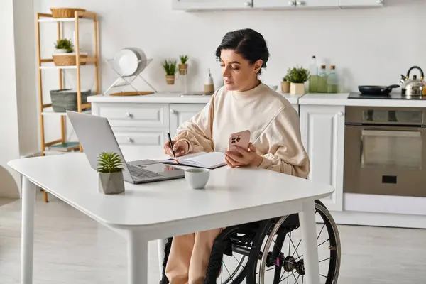 Eine behinderte Frau im Rollstuhl mit einem Laptop am Küchentisch, die aus der Ferne zielstrebig und konzentriert arbeitet. — Stockfoto
