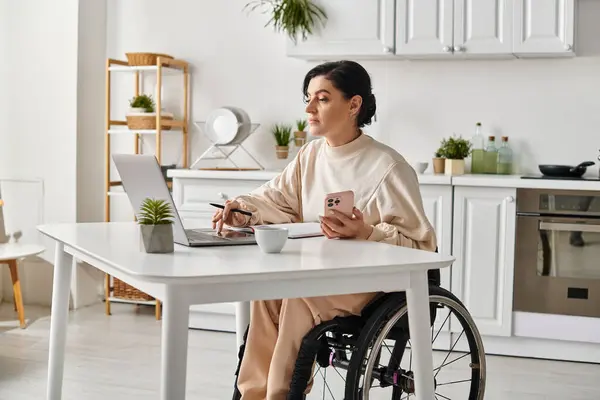 Une femme handicapée dans un fauteuil roulant utilisant un ordinateur portable à la maison dans sa cuisine pour le travail à distance. — Photo de stock