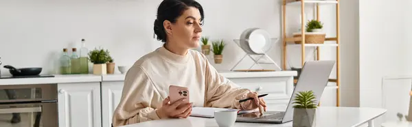 Женщина за кухонным столом, сосредоточенная на ноутбуке и мобильном телефоне. — стоковое фото
