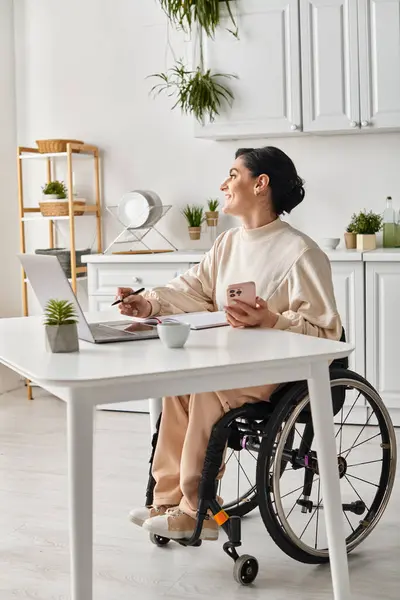 Une femme handicapée dans un fauteuil roulant travaillant sur un ordinateur portable dans sa cuisine — Photo de stock