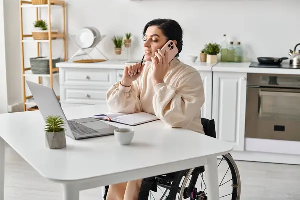 Una mujer discapacitada en una silla de ruedas multitarea, trabajando a distancia, y charlando en un teléfono celular en su cocina. - foto de stock