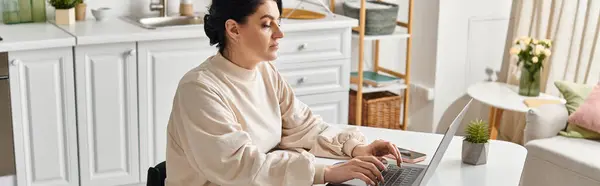 Uma mulher deficiente em uma cadeira de rodas, trabalhando remotamente, senta-se em uma mesa usando um computador portátil em sua cozinha. — Fotografia de Stock