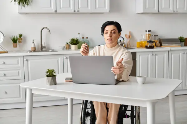 Жінка-інвалід у інвалідному візку зосереджена на ноутбуці за кухонним столом, займаючись віддаленою роботою або дозвіллям. — стокове фото