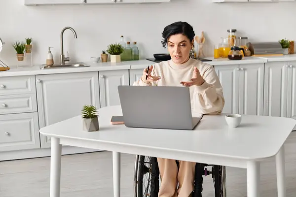 Uma mulher deficiente em uma cadeira de rodas sentada em uma mesa de cozinha, focada em seu computador portátil. Ela está a trabalhar remotamente.. — Fotografia de Stock