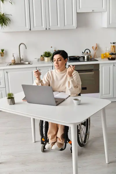 Une femme handicapée en fauteuil roulant travaille à distance dans sa cuisine, se concentrant sur un ordinateur portable sur la table. — Photo de stock