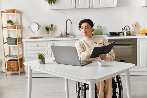 Uma mulher deficiente em uma cadeira de rodas senta-se em uma mesa de cozinha, trabalhando em seu laptop com determinação e foco. — Fotografia de Stock