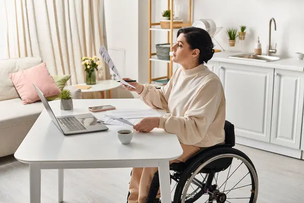 Женщина-инвалид в инвалидной коляске, работающая на ноутбуке на кухне, демонстрирующая расширение возможностей и технологический прогресс. — стоковое фото
