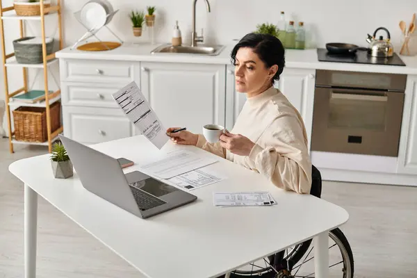 Una mujer en silla de ruedas trabaja en su portátil, rodeada de papeles, en una acogedora cocina. - foto de stock