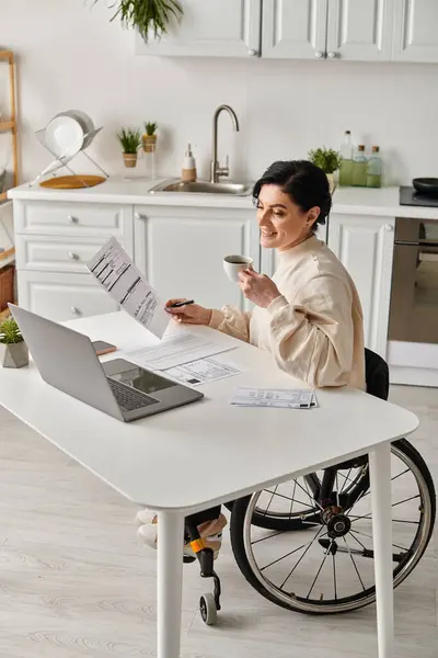Uma mulher em uma cadeira de rodas está sentada em uma mesa em sua cozinha, focada em trabalhar em seu laptop remotamente. — Fotografia de Stock