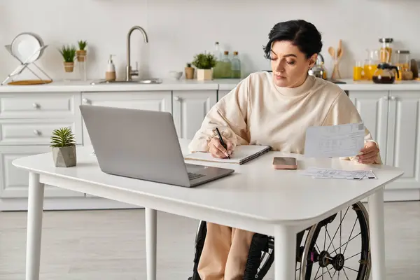 Une femme handicapée dans un fauteuil roulant assise à une table avec un ordinateur portable, travaillant à distance dans sa cuisine. — Photo de stock