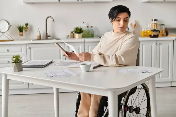 Una mujer discapacitada en una silla de ruedas trabaja a distancia usando una tableta en su mesa de la cocina. - foto de stock