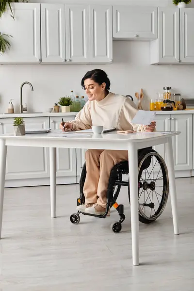 Uma mulher deficiente em uma cadeira de rodas está trabalhando em uma mesa de cozinha. — Fotografia de Stock