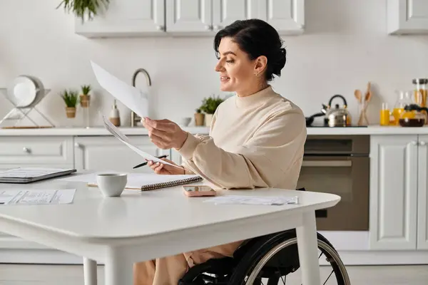 A disabled woman in a wheelchair sits in her kitchen, holding a piece of paper while working remotely. — Stock Photo