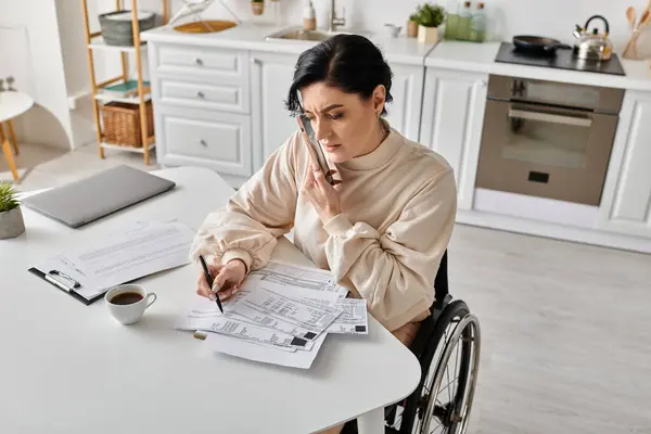 A disabled woman in a wheelchair reads a paper while working remotely from her kitchen. - foto de stock