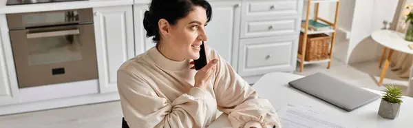 A brunette woman talks at phone and working remotely at a table in her kitchen. — Stock Photo