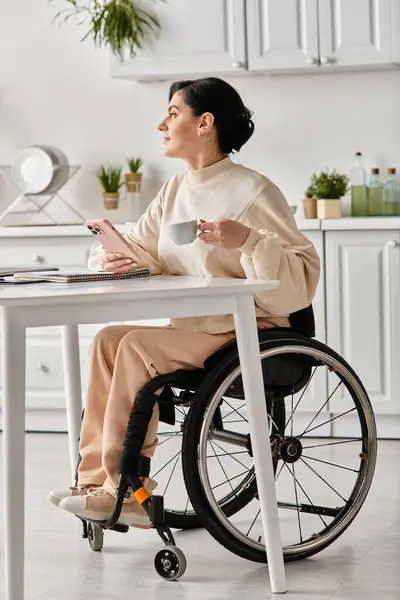 Une femme en fauteuil roulant travaillant sur un ordinateur portable à une table dans sa cuisine, faisant preuve de détermination et de concentration. — Photo de stock
