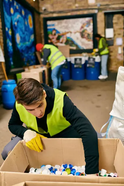 Young eco-conscious volunteer in yellow gloves and vest cleaning a cardboard box with dedication. — Stock Photo