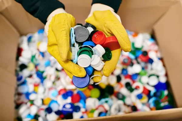 A young volunteer in yellow gloves carefully holding a bunch of colorful bottle caps while sorting trash with eco-conscious peers. — Stock Photo