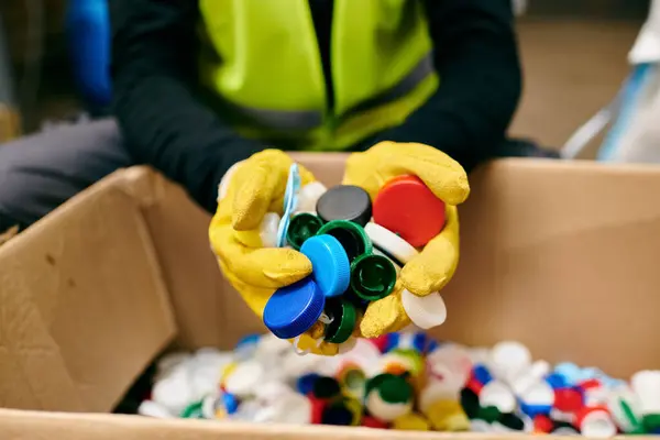 A young volunteer in yellow gloves carefully holding a bunch of colorful bottle caps while sorting trash — стокове фото