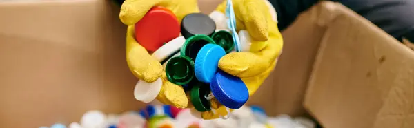 A young volunteer in yellow gloves holding a bunch of colorful bottle caps while sorting waste — стокове фото