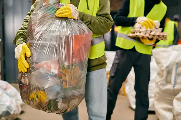 Two young volunteers in safety vests, gloves, and holding a huge bag of garbage while sorting trash. — Foto stock