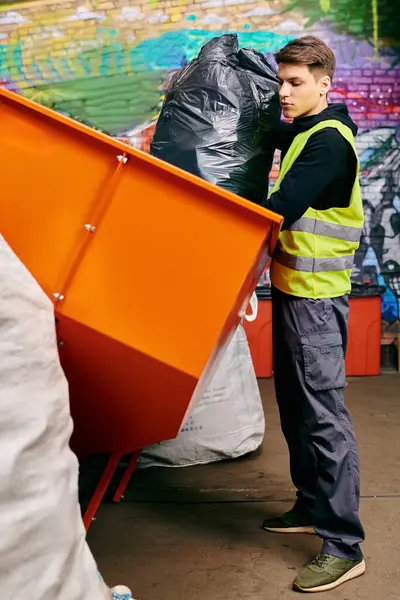 Young volunteer in gloves and safety vest stands next to dumpster, sorting trash in front of a wall. — Fotografia de Stock